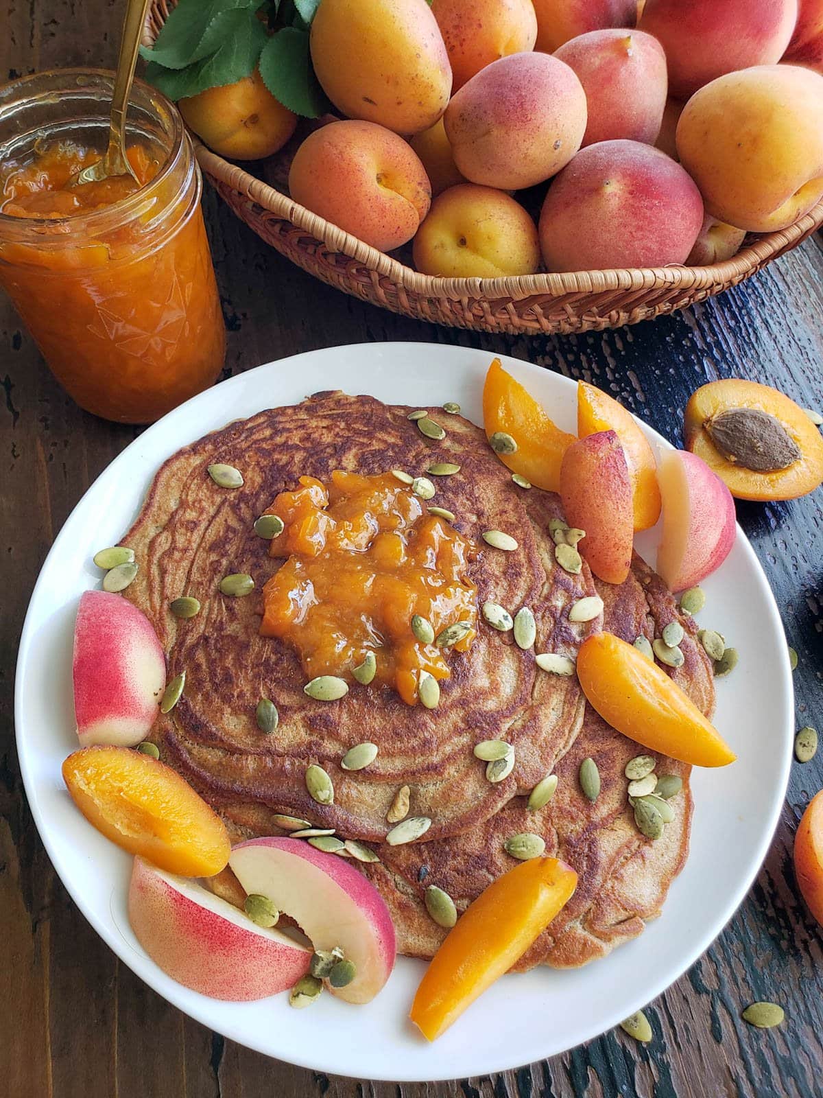 Cooked flapjacks on a white ceramic plate have been topped with fresh slices of apricot and white peach, pumpkin seeds, and a dollop of apricot jam. 