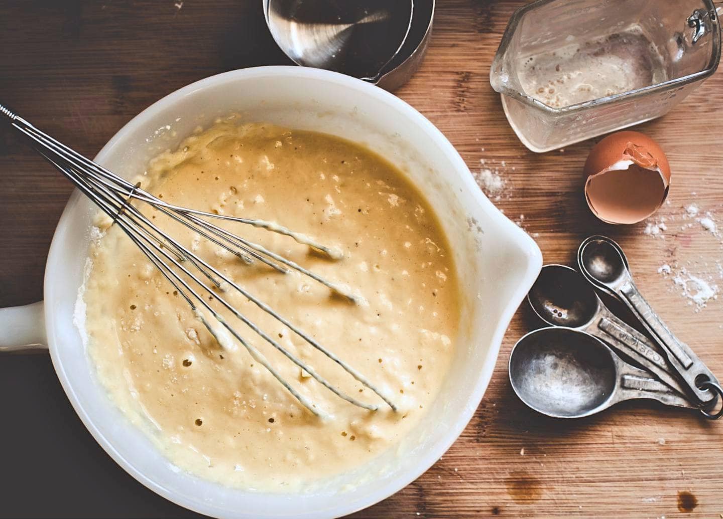A white mixing bowl is full of sourdough pancake batter with a metal whisk resting inside it. Around the bowl are tablespoon measurements, an egg shell, and a few soiled measuring cups. 