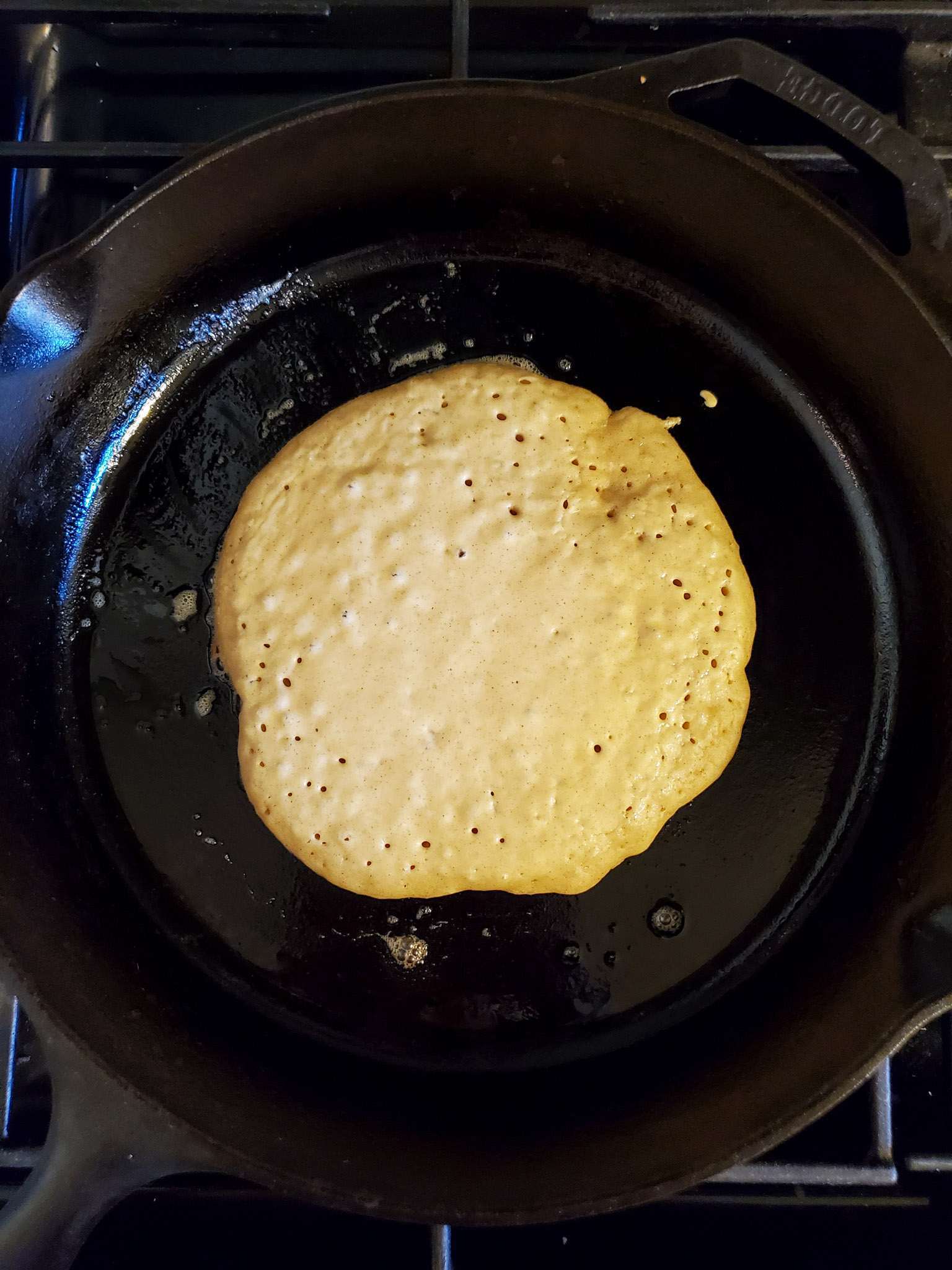 Sourdough pancake batter freshly poured into the middle of a cast iron skillet. The melted butter streaks are visible in the pan, there are bubbles forming throughout the pancake and the edges are starting to firm up and turn slightly brown. 