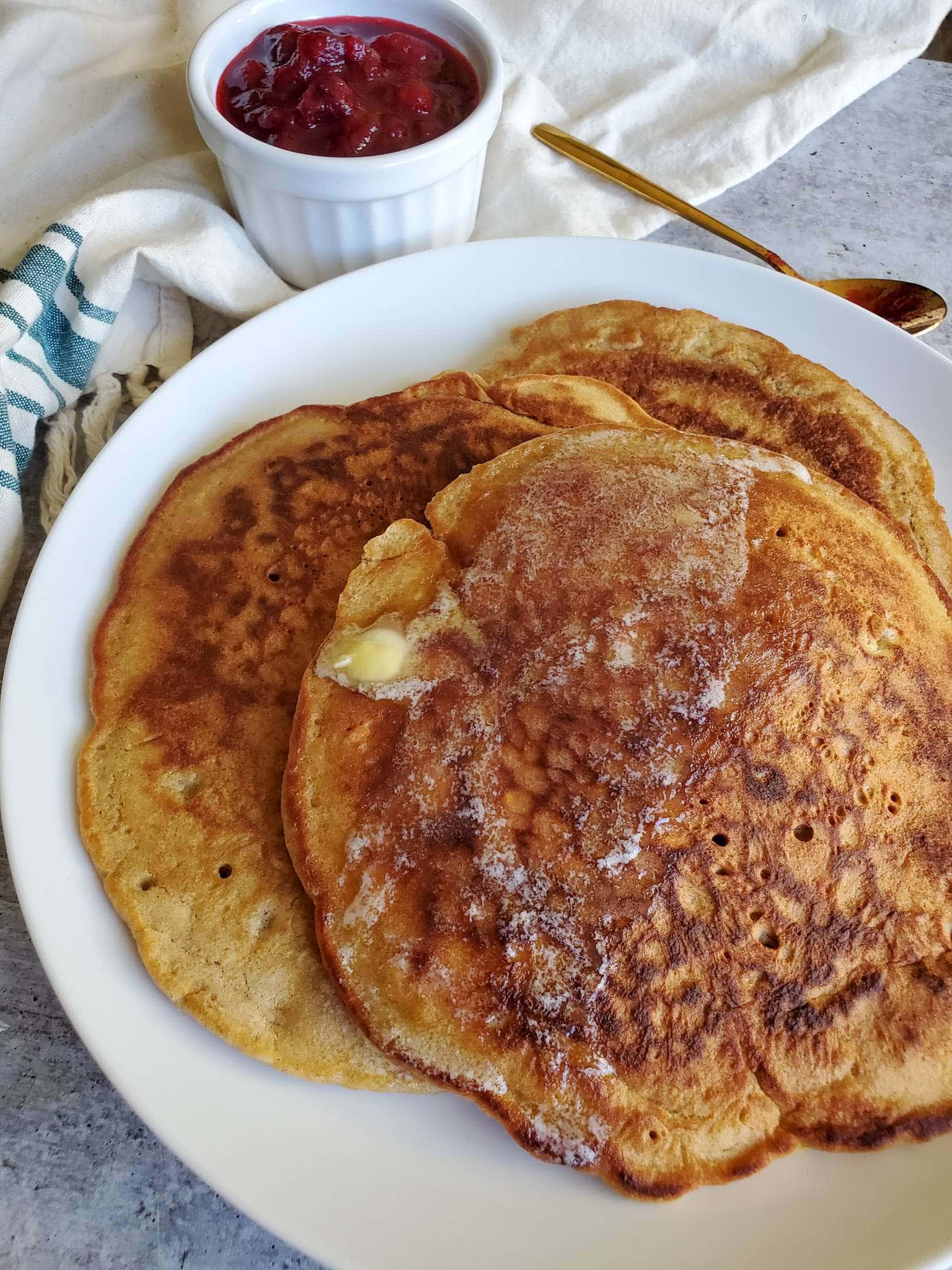 A stack of flapjacks sit atop a white ceramic plate. A pad of butter is sitting on the top pancake and is shrinking in size as it melts throughout the top portion of pancake. A ramekin of fresh cranberry sauce sits in the background next to a gold spoon. 