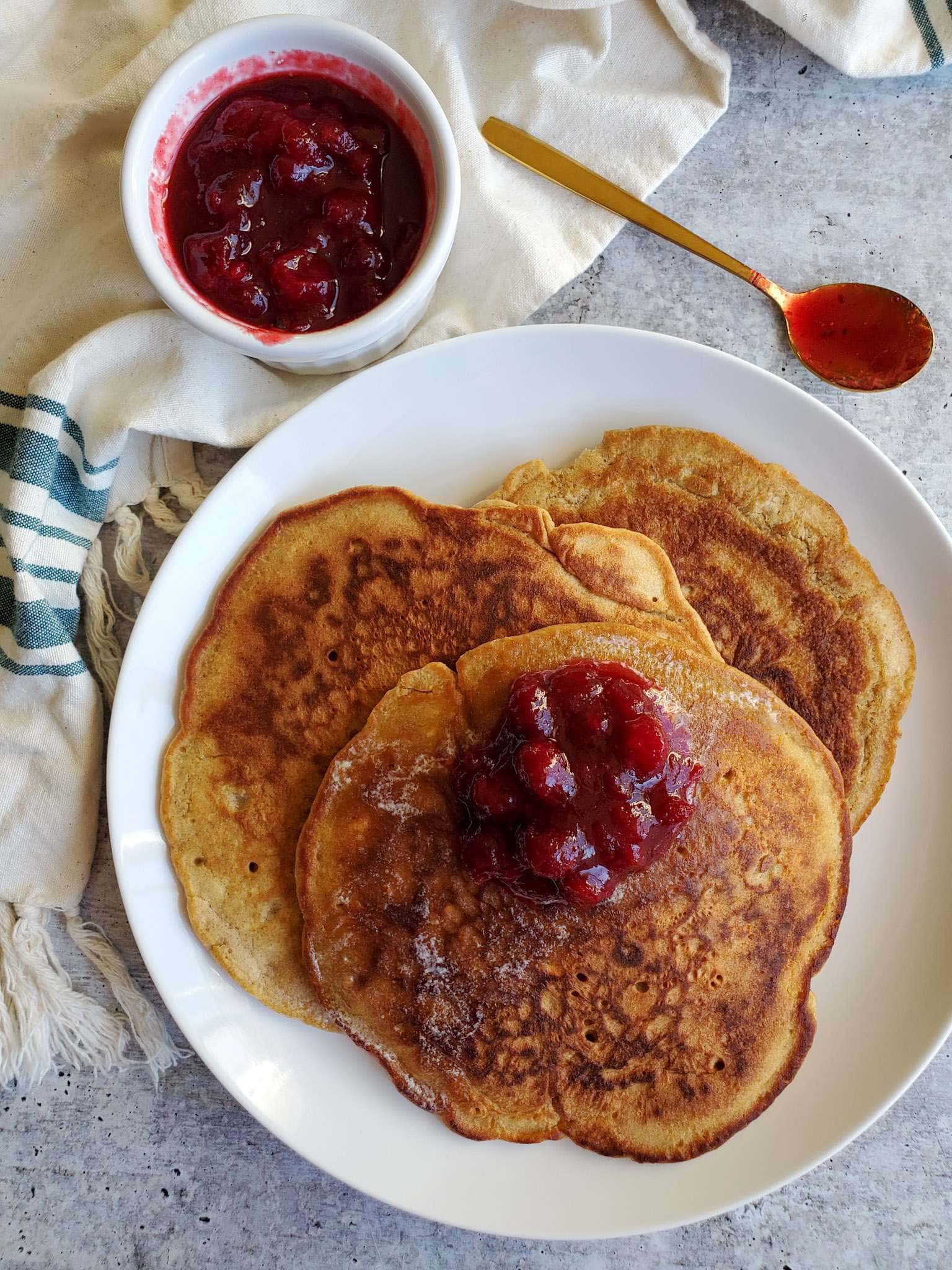 A stack of pancakes sit atop a white ceramic plate. A dollop of cranberry sauce sits on the top pancake.  A ramekin of fresh cranberry sauce sits in the background next to a gold spoon that is now red from the sauce. 