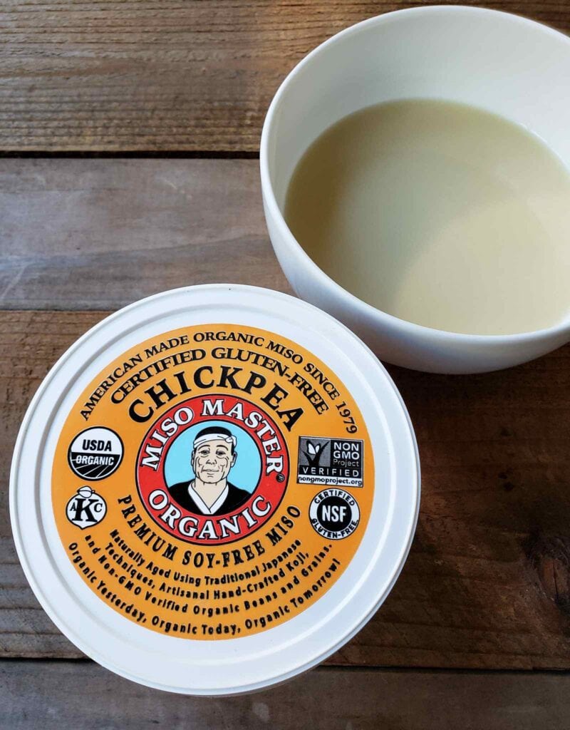 A container of Miso Master organic chickpea miso paste sits next to a white ceramic bowl of miso broth that is a creamy milky color. A slatted wooden backdrop is the background.