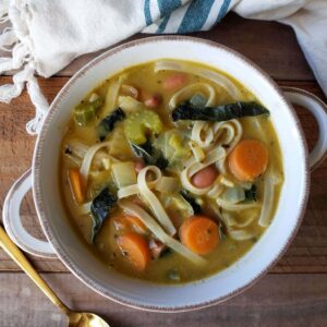 A white ceramic bowl with handles is full of vegetarian chicken noodle soup, carrots and greens are amongst the noddles and broth.