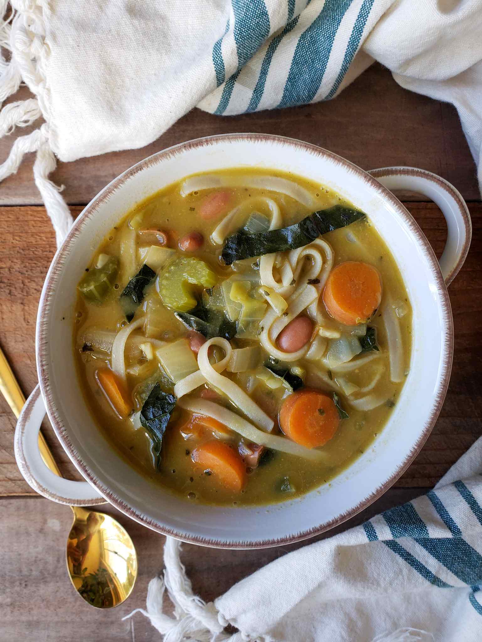 Looking down on a bowl of vegan no-chicken noodle soup inside a white ceramic bowl with two handles. A golden spoon is off to the side next to a white cloth towel with a few blue lines running widthwise across it. Carrots, celery, kale, onion, and noodles are visible in the yellowish lighter brown broth of the soup.