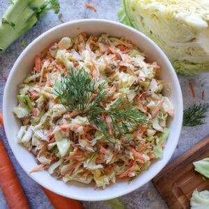 A white ceramic bowl is full of cabbage and carrot coleslaw. Fresh cabbage and carrots surround the bowl.