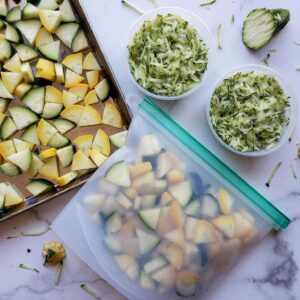 A baking sheet contains raw summer squash that has been cut into bite sized pieces. A silicone bag is next to it filled with the same type and cut of squash. Two small plastic containers are filled with grated summer squash.