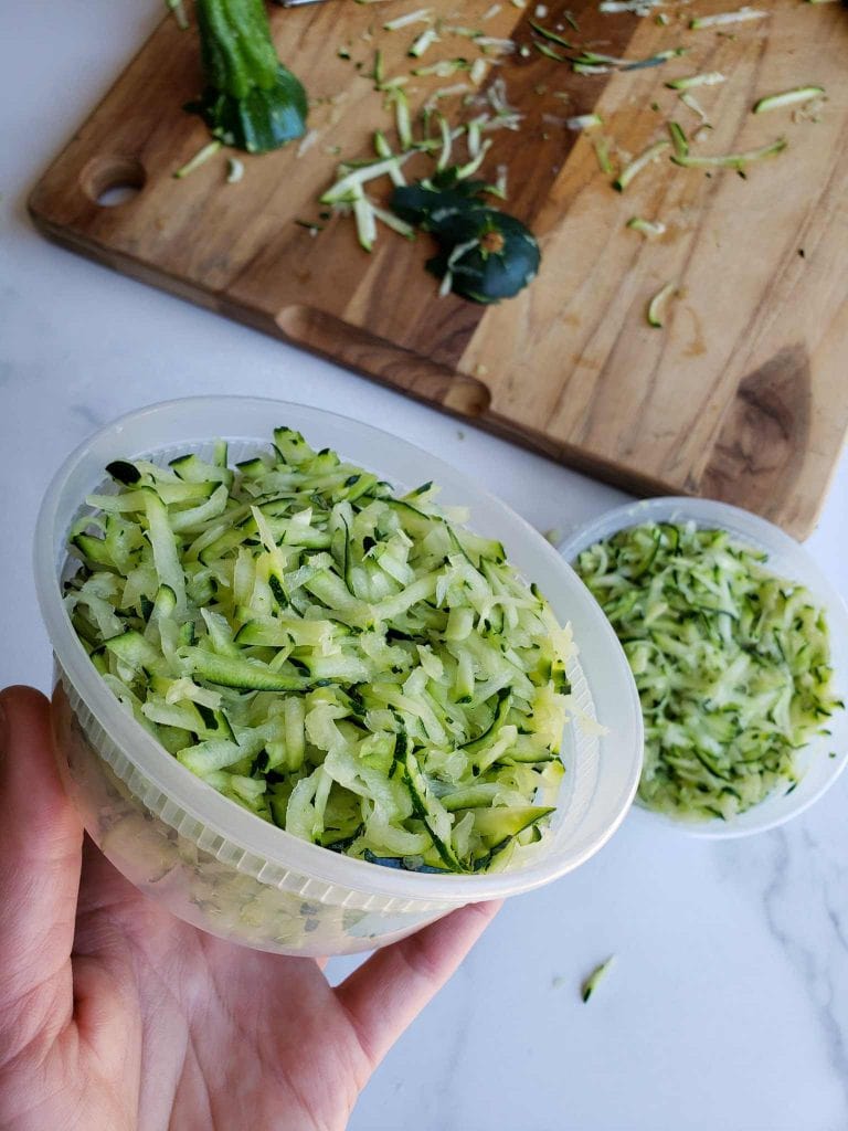 A BPA free cup container is shown full of shredded zucchini. Below lies another container full along with a cutting board that contains the stem end and butt end of the squash along with some remaining shreds of zucchini. 