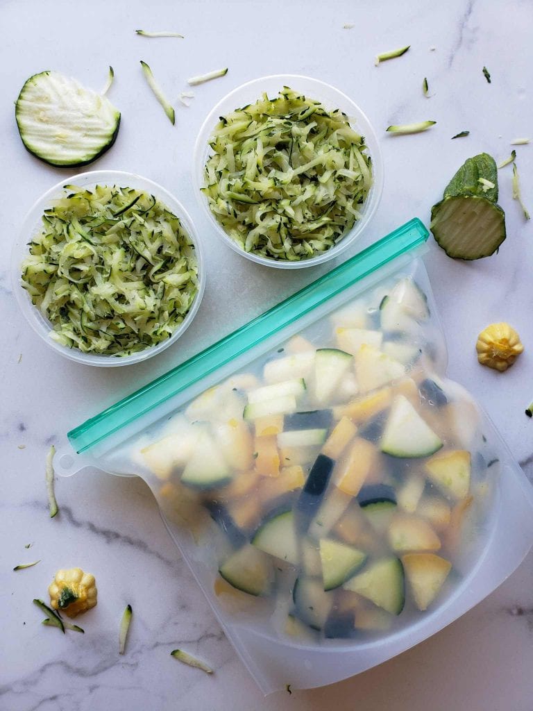 A flat lay image containing two 8 oz. BPA free containers full of shredded squash. Below lies the silicon bag full of squash chunks. Accenting the prepared squash are random grates of zucchini, slices of zucchini, as well as stem pieces from the squash. 