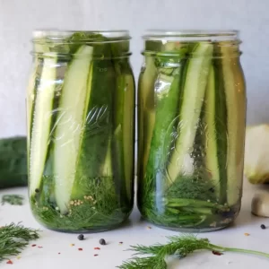 Two quart mason jars are filled with long spears of pickles. Surrounding the jars are onion, cucumber, fresh dill, and peppercorns.