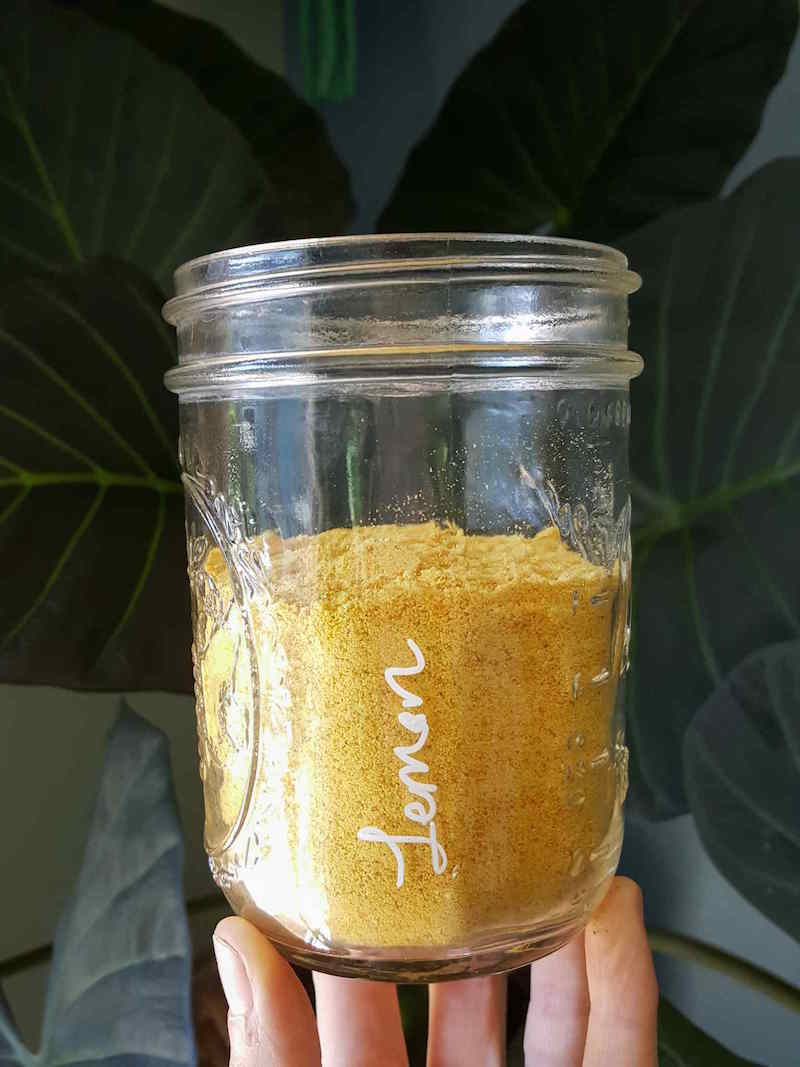 A hand is holding a pint mason jar up to display the golden orange lemon powder that resides within. The jar is two thirds of the way full and the jar is marked "lemon" with a wine glass marker.