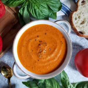 A white bowl with handles is full of tomato soup. Surrounding the bowl are fresh tomatoes, basil leaves, and a slice of bread.