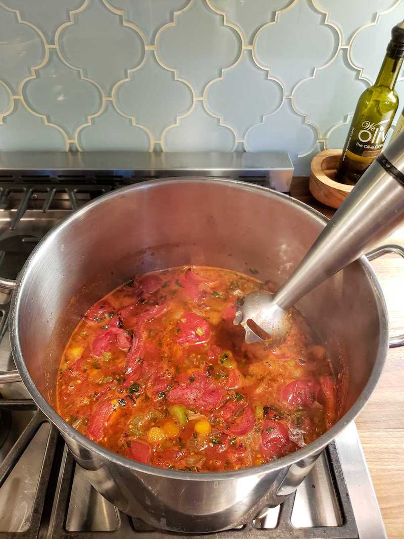 A three gallon stainless steel stock pot full of the soup ingredients which contain tomatoes, celery, carrots, onions, red bell pepper, vegetable broth, coconut milk, and various spices. An immersion blender is poised over the surface of the soup, ready to blend the mixture into a smooth texture.