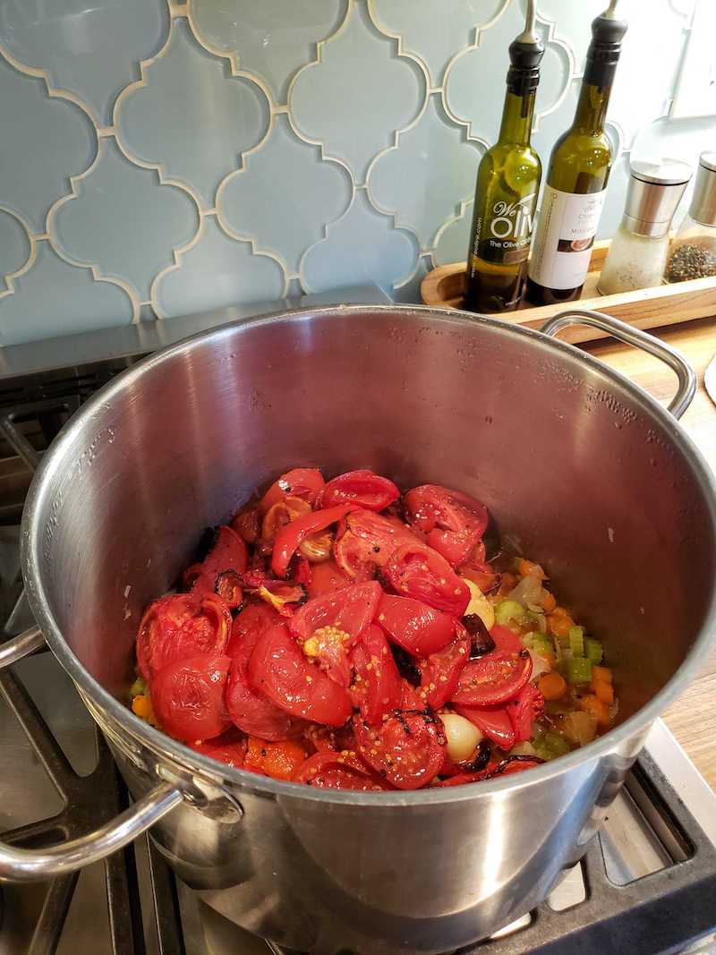 A three gallon stainless steel stock pot has roasted tomatoes, red bell peppers, and garlic are piled atop celery, carrots, and onions as the base for the fresh roasted tomato basil soup.
