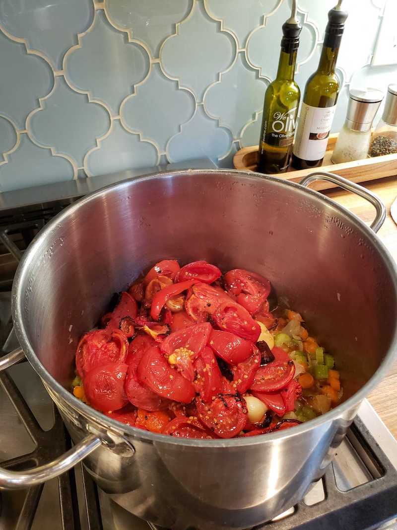 A three gallon stainless steel stock pot has roasted tomatoes, red bell peppers, and garlic are piled atop celery, carrots, and onions as the base for the fresh roasted tomato basil soup. 