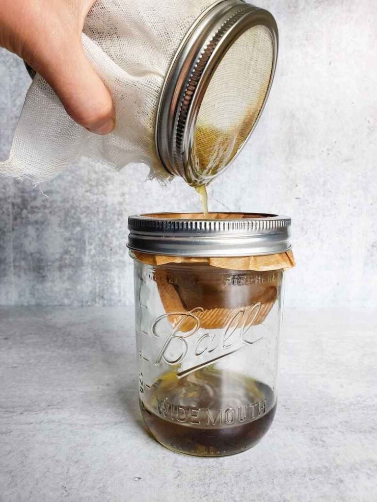 A pint mason jar with a cheesecloth attached to the rim of the jar is pouring liquid into another pint mason jar with a coffee filter around the rim of the jar. There is some golden liquid at the bottom of the jar with the filter.