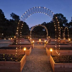 A raised bed garden at dusk, there are arch trellises on a number of the beds which are lined with twinkle lights which are lighting up the area. Solar powered tiki torches are also set in various raised beds throughout the space.