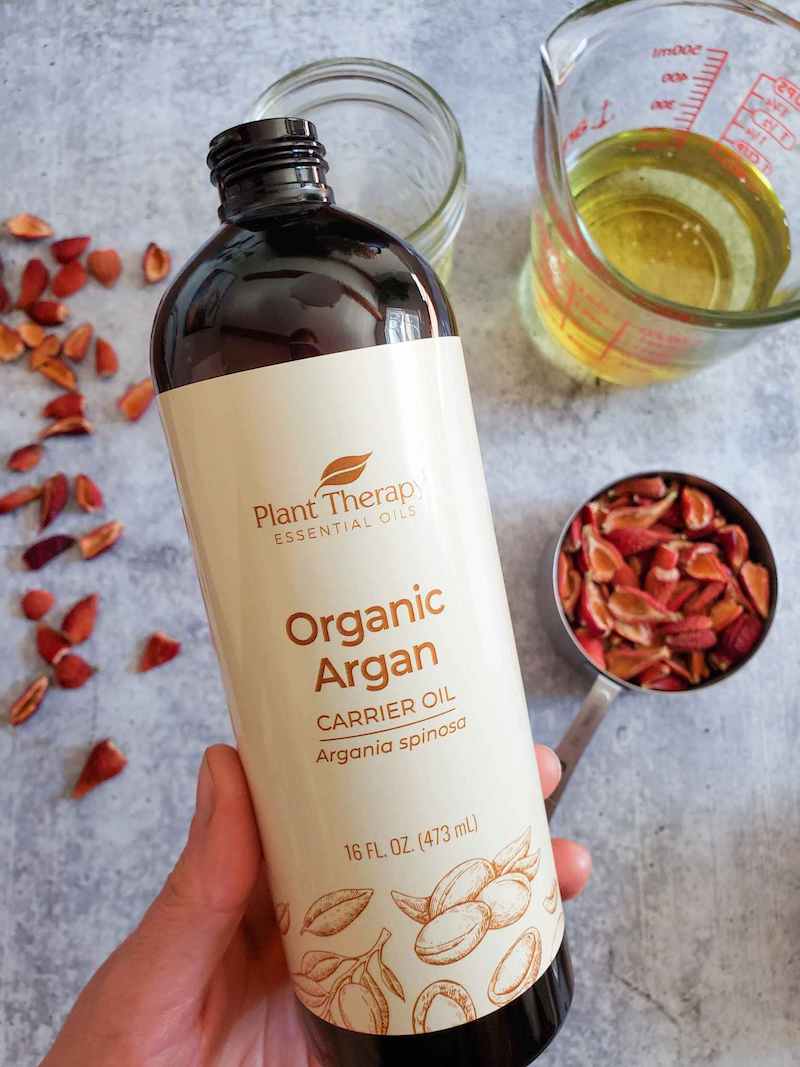 DeannaCat's hand is holding a bottle of organic argan oil. Below lies a stainless steel measuring cup full of dried rosehips as well as a liquid glass measuring cup and an empty pint mason jar. There are more dried rosehips scattered along the left edge of the image, covering some would be empty space. 