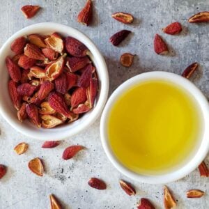 A white ramekin is filled with rose hip oil while another ramekin is full of dried rose hips, more dried rosehips are scattered around the area surrounding the ramekins.
