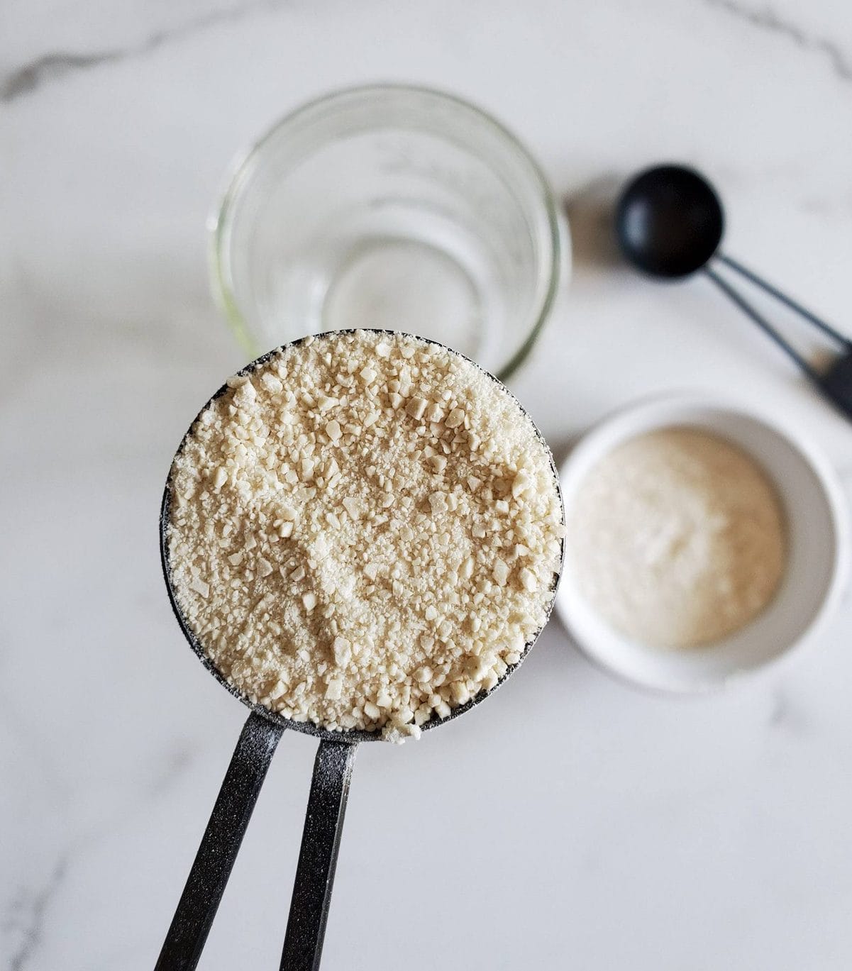 A half tablespoon measurement is full  of dried sourdough starter which has been blended into a crumbly powder. Below lies a ramekin of flour and a mason jar.