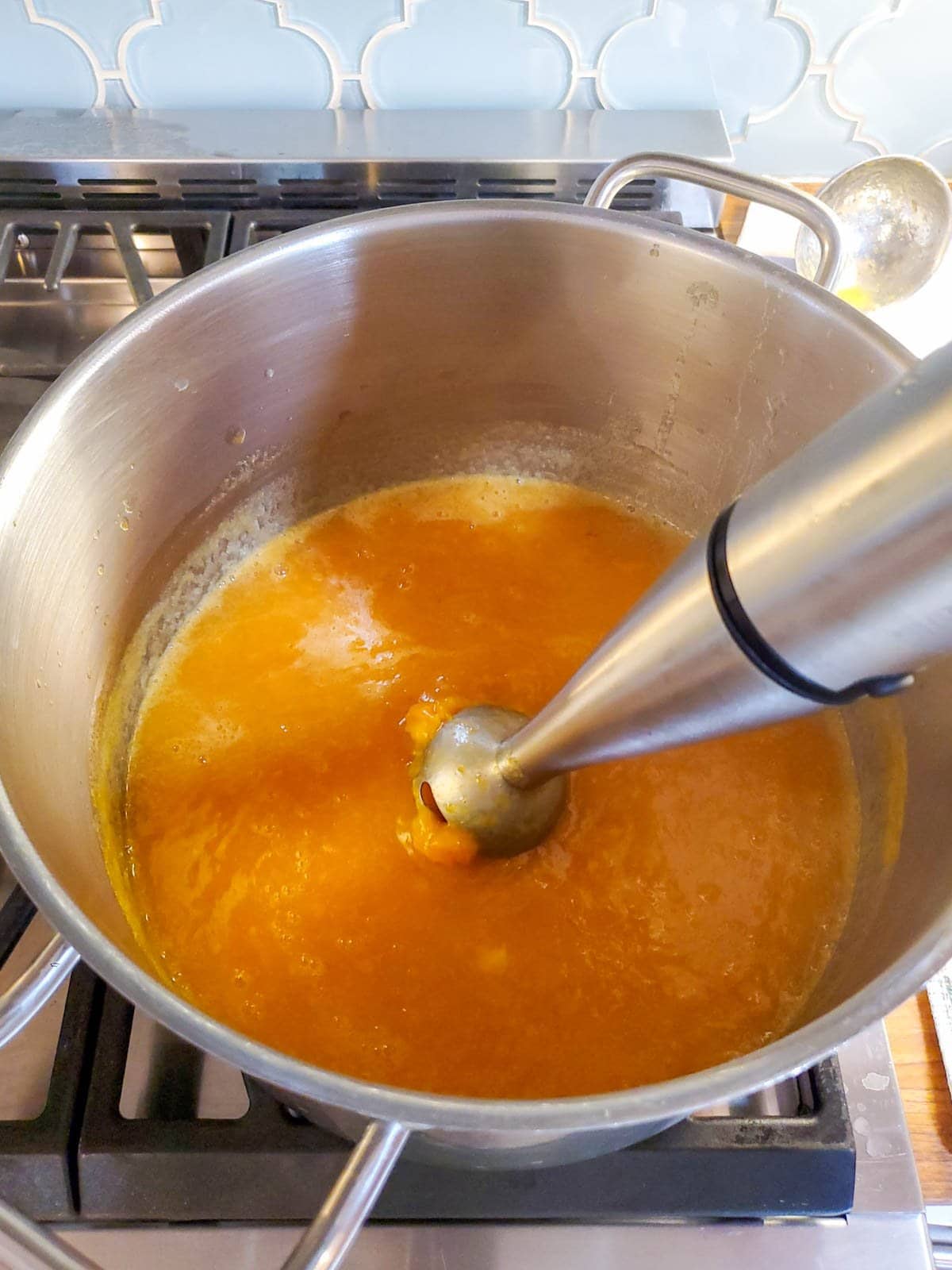 A handheld immersion blender is poised inside a large stainless steel pot with bright orange peach jam cooking in the bottom of the pan, helping to thicken and blend it smooth