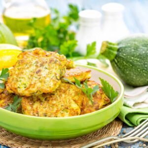 A dish of zucchini fritters stacked high are surrounded by a few fresh squash.