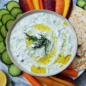 A bowl of yogurt dill sauce with a drizzle of olive oil with a sprig of dill in the middle of it. Surrounding the bowl are cucumber slices, carrots sticks, and crackers.