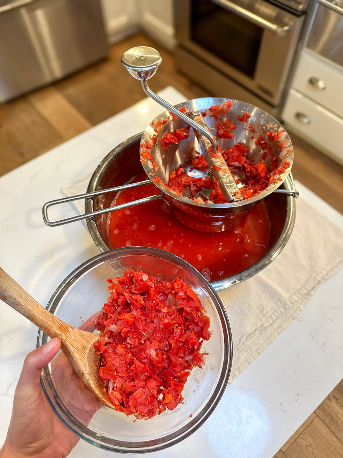 A hand holding a small bowl of saved tomato skins, with a larger bowl of strained tomato juice with a food mill sitting on top in the background