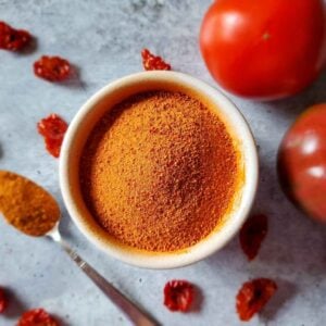A white ramekin is full of dried tomato powder which is bright red in color. A few fresh tomatoes are arranged around the bowl along with a few dried tomato pieces. A spoon is resting next to the ramekin with a heaping spoonful of tomato powder.
