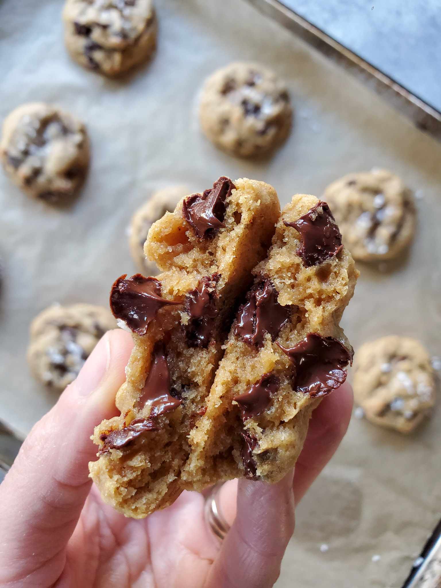 A close up image of a cookie that has been pulled apart in two just after baking. The two halves are sandwiched together to reveal the gooey insides from the melty chocolate chips. A baking sheet full of cookies resides below in the background. 