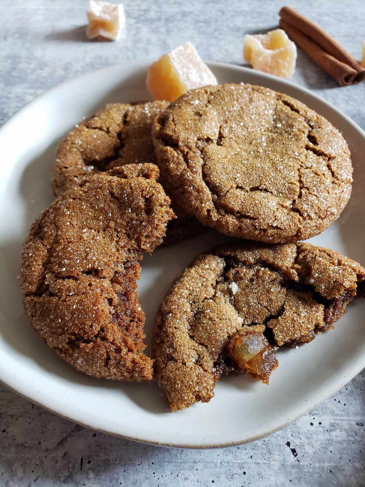 Three sourdough ginger molasses cookies stacked on a white ceramic plate. One of them has been torn in half, one half propped up on the other two. There are crystalized ginger chunks and a cinnamon stick in the background behind the cookies. Use starter discard to make many baked good treats.
