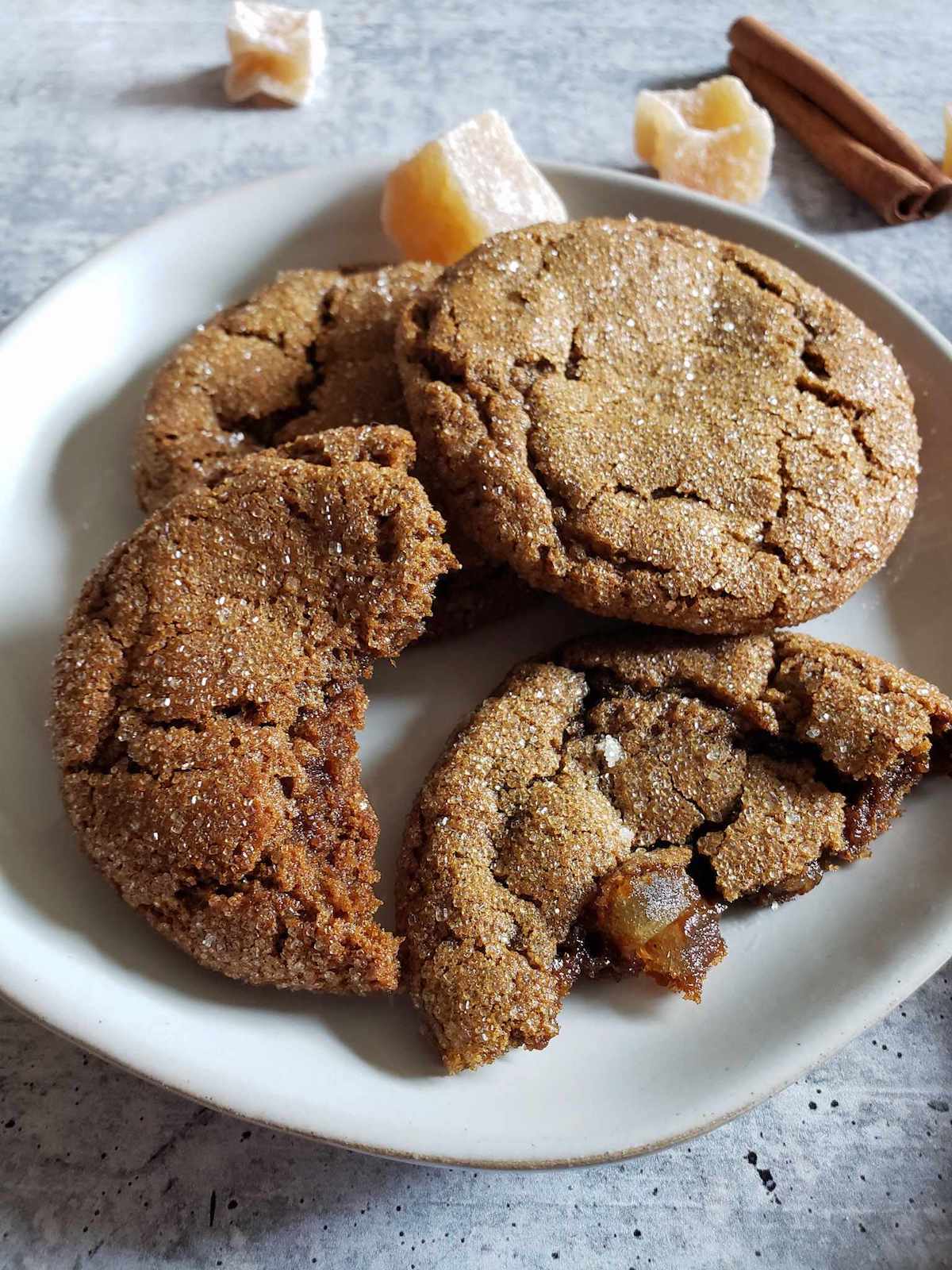 Three sourdough ginger molasses cookies stacked on a white ceramic plate. One of them has been torn in half, one half propped up on the other two. There are crystalized ginger chunks and a cinnamon stick in the background behind the cookies.  Use starter discard to make many baked good treats.