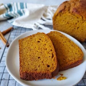 Two slices of sourdough pumpkin bread are arranged on a small white plate. The remaining loaf of bread is sitting just beyond on a wire cooling rack.