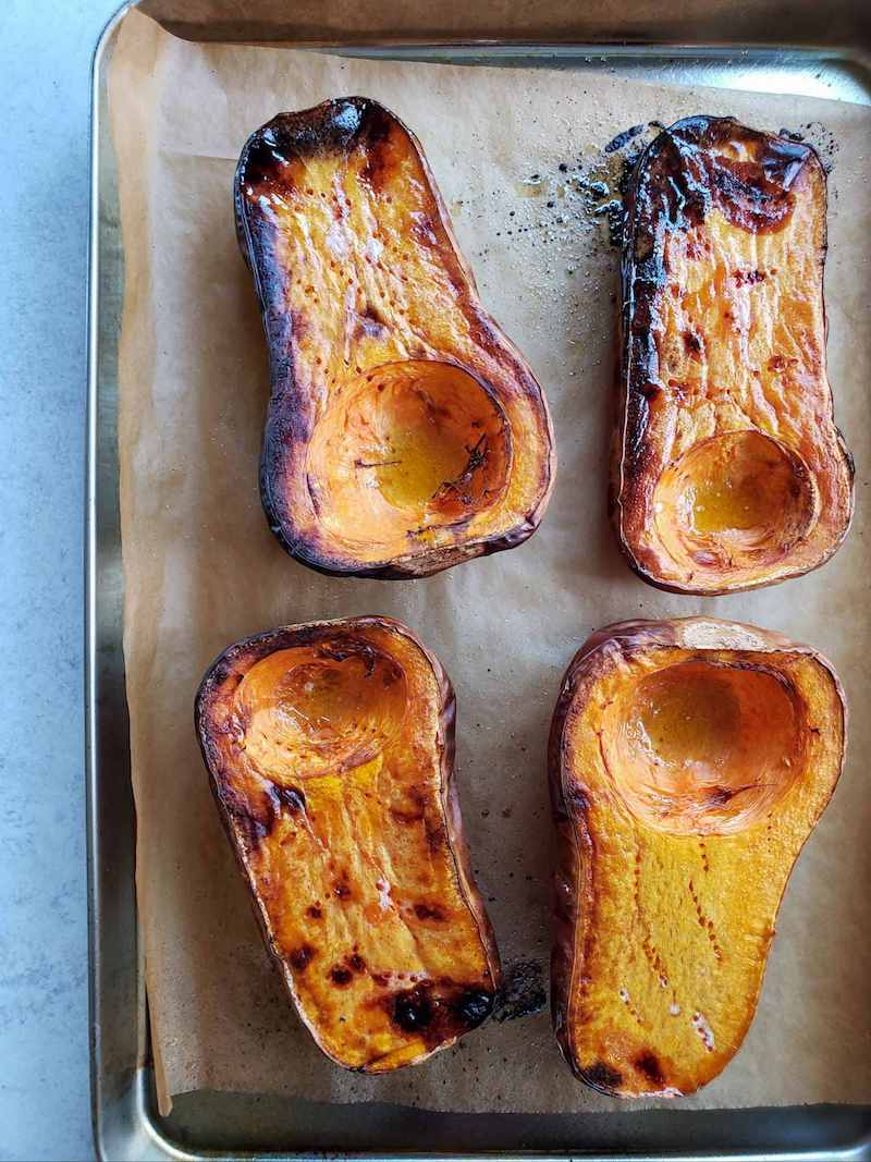 Four halves of butternut squash sit atop a baking sheet lined with parchment paper after being roasted in the oven. Bits of dark brown and black are evident on the surface after caramelizing.