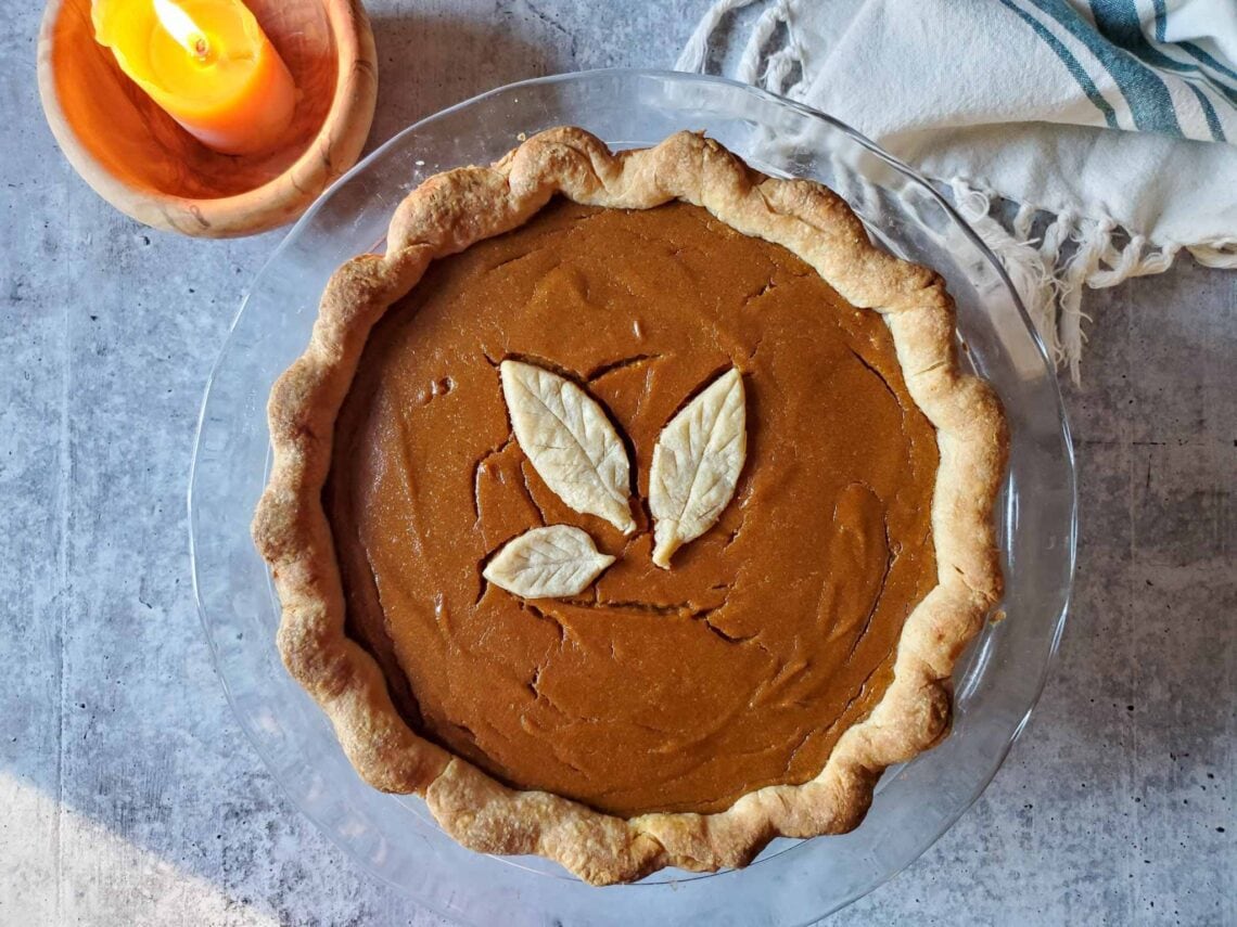 A freshly baked butternut squash pumpkin pie is in a glass pie baking dish. A leaf made out of dough is in the middle of the pumpkin pie.