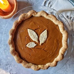 A freshly baked butternut squash pumpkin pie is in a glass pie baking dish. A leaf made out of dough is in the middle of the pumpkin pie.