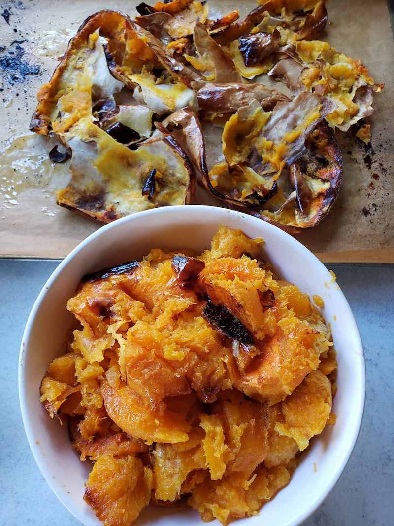 A white ceramic bowl full of roasted squash flesh. There is parchment paper on a baking sheet that contain the skin from the squash.