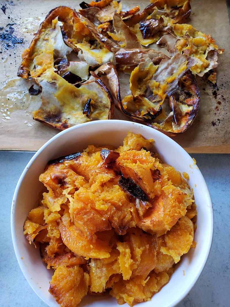 A white ceramic bowl full of roasted squash flesh. There is parchment paper on a baking sheet that contain the skin from the squash. 