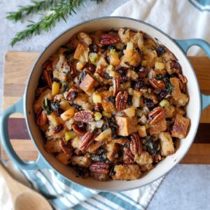 A two handled ceramic pot is full of vegetarian sourdough stuffing, pieces of cranberry, apple, and greens mixed in with the larger pieces of sourdough bread.