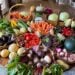 A round table contains a huge harvest of fresh vegetable varieties, from melons, eggplant, basil, tomatoes, carrots, turnips, hard squash, cabbage, grapes and summer squash.
