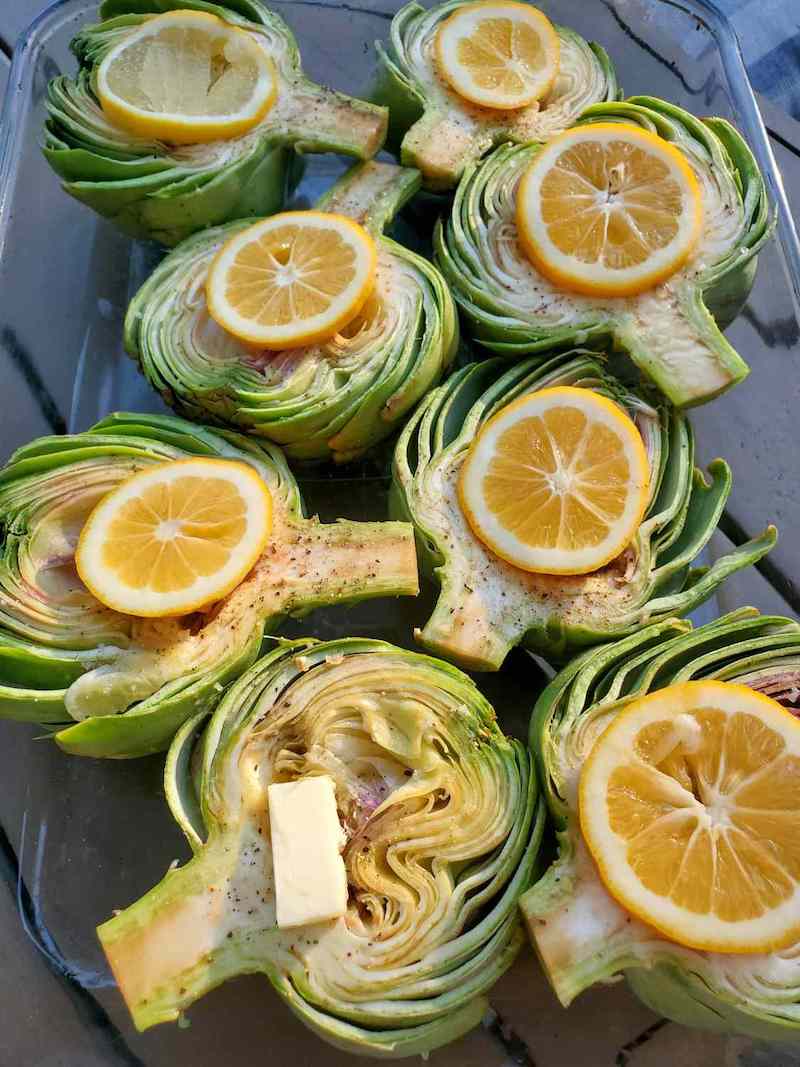 Beautiful green globes have been cut in half, sitting face up in a glass baking dish with a dollop of butter and a slice of lemon laid over the top of each one.