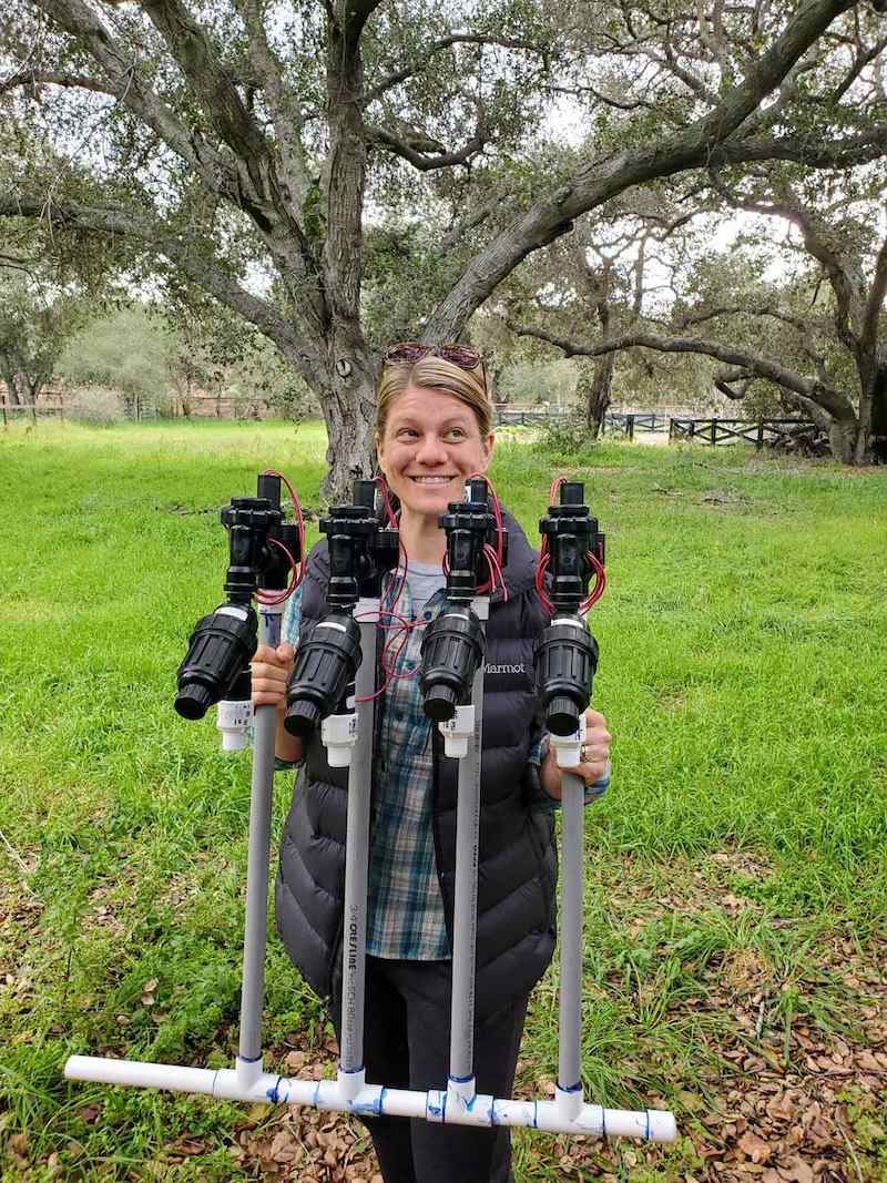 DeannaCat is holding a 4-station valve manifold with mainline PVC attached to the bottom. This section will be connected to the end of a preexisting water line.