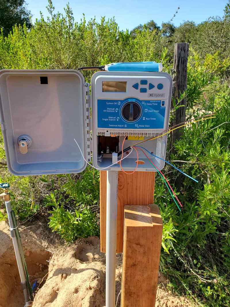 An irrigation control box is connected to a 2x6 section of wood connected to a 4x4 post. The controller is open and the wires from the valves are splayed out inside the box, they need to be inserted into the corresponding zone sections inside the controller. 