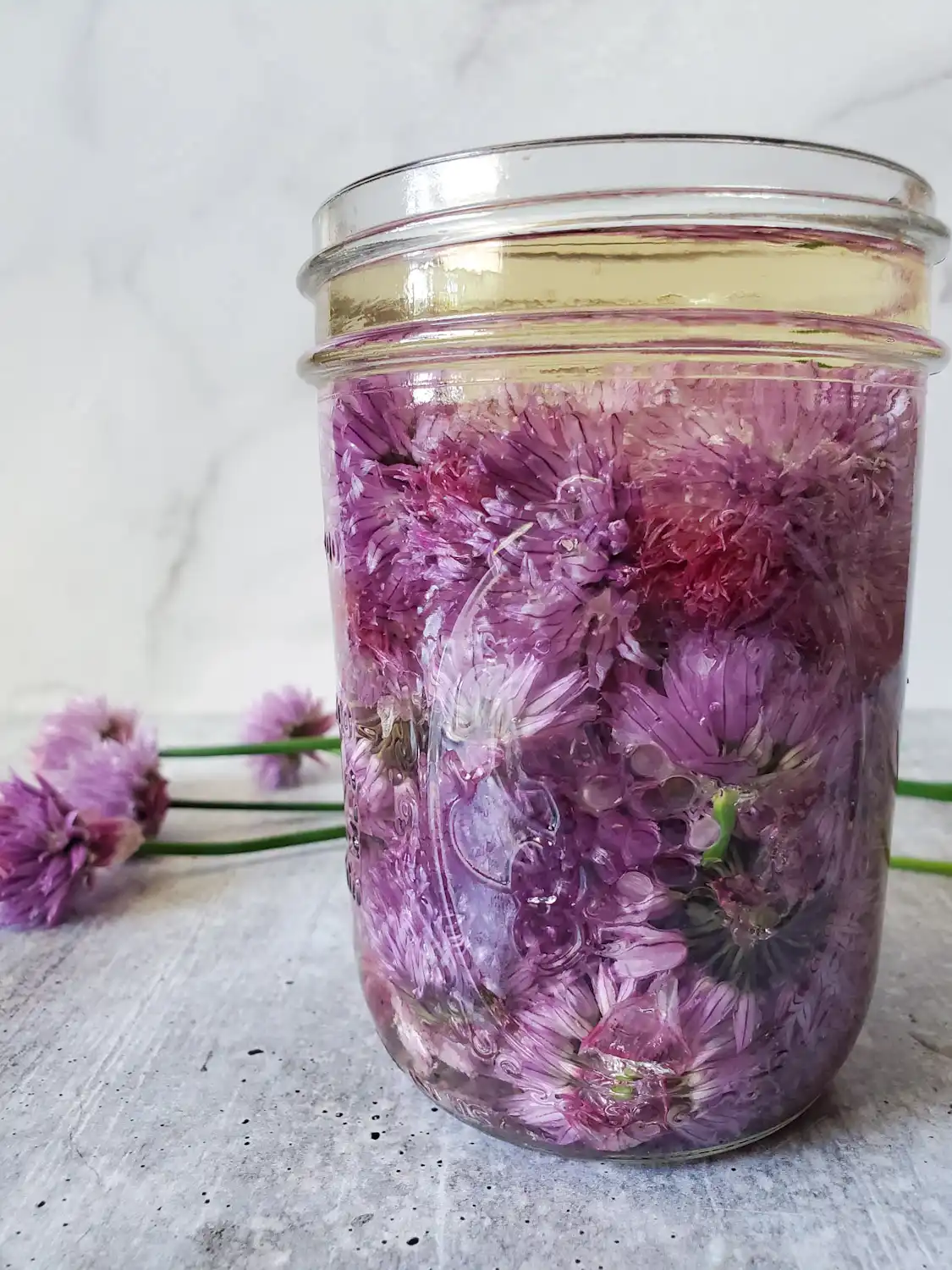 A pint mason jar full of chive blossoms that are covered completely in vinegar. Beyond are a few more chives with light purple blossom blooms atop the green chive.