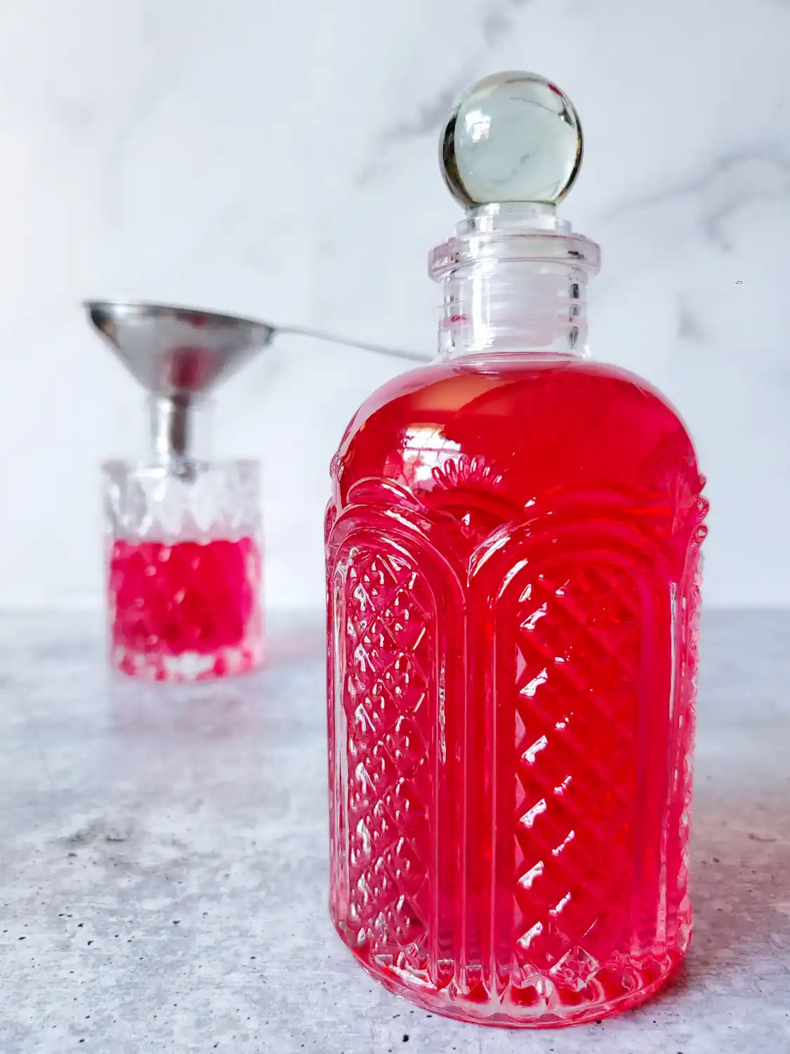A fancy glass bottle is in the foreground full of brilliantly light red liquid. Beyond there is a smaller bottle that is halfway full of the same liquid, a stainless steel funnel is sitting inside the top opening.