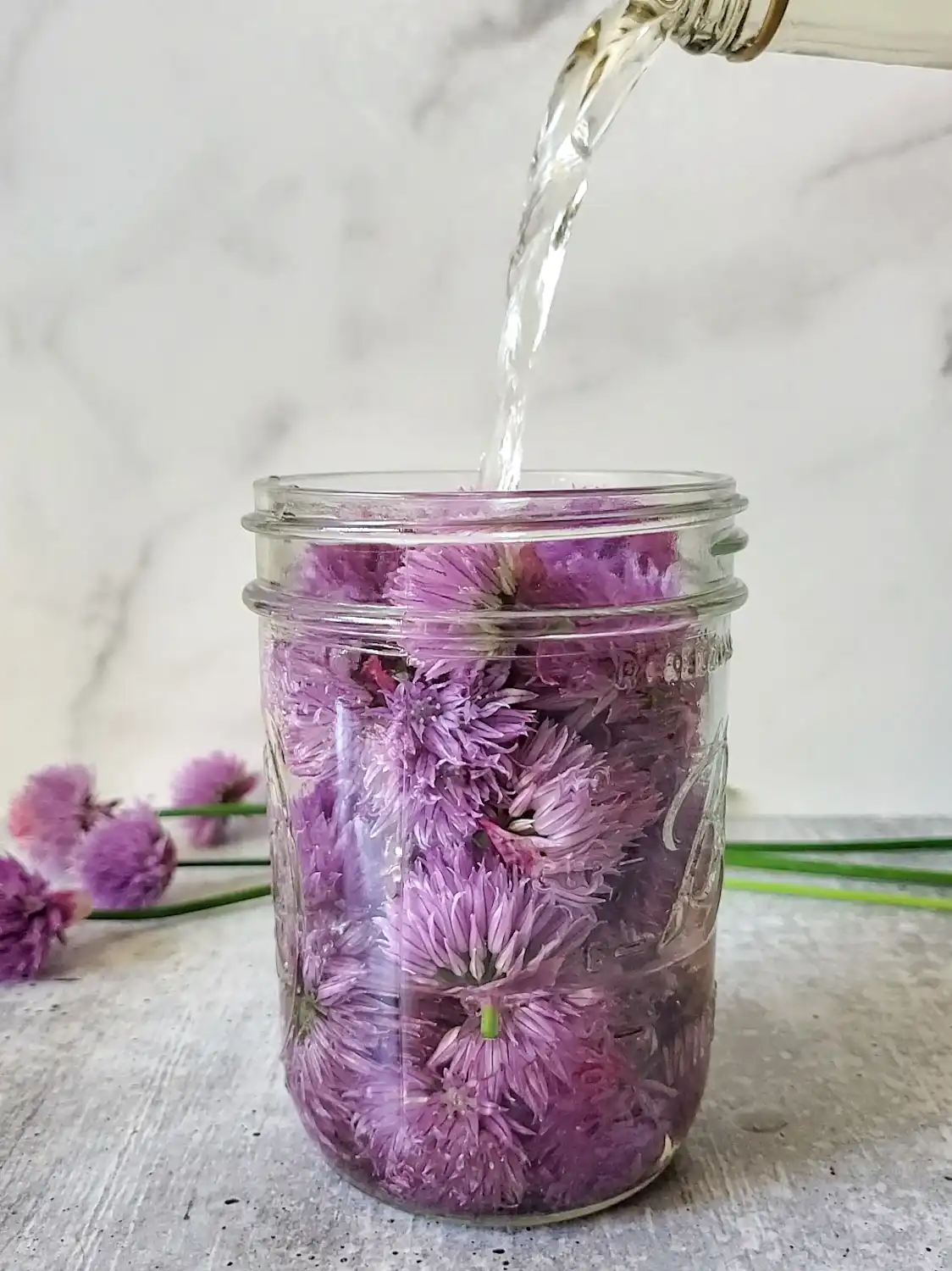 A stream of clear liquid is streaming down from the top of the image into a pint mason jar full of purple flower blooms.