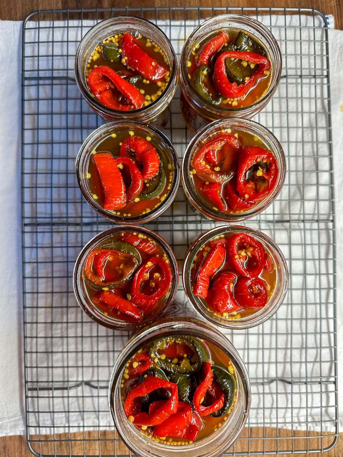 Looking down over 7 canning jars on a cooling rack ful of red jalapeno rings in syrup