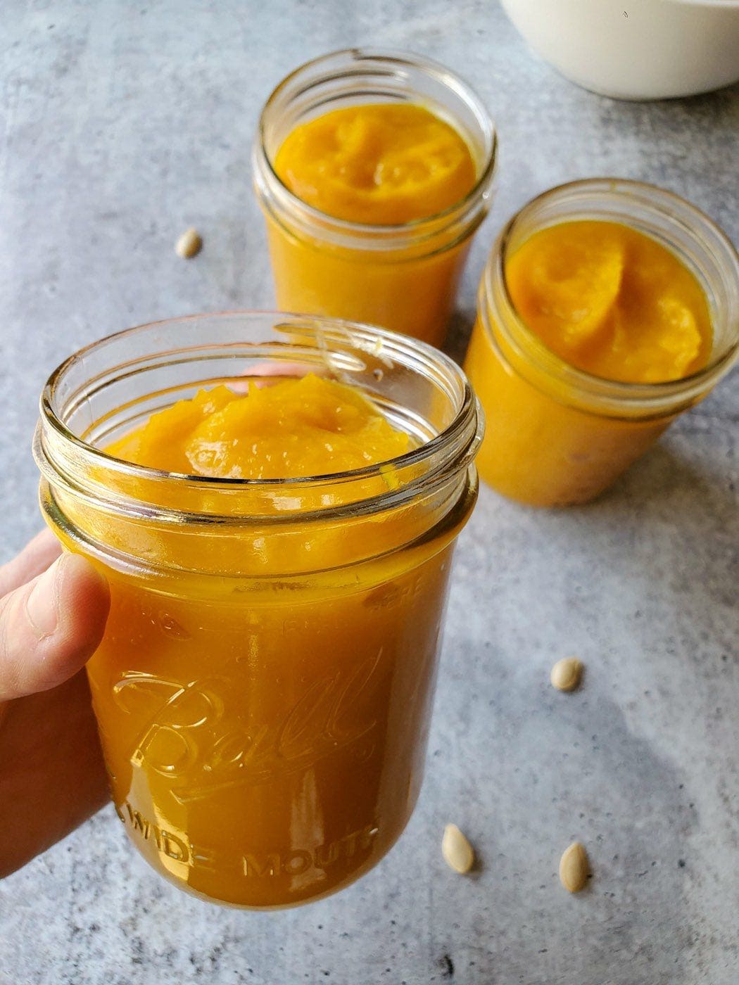 A hand is holding a pint mason jar full of homemade pumpkin puree. It is vibrant orangish yellow in color, in the background there are two more pints of the pumpkin puree along with a few pumpkin seeds scattered about.