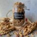 A flip top jar is overflowing with sourdough granola, the jar has a black label on it that reads "sourdough granola". There are chunks of sourdough granola surrounding the jar along with a wooden scoop which contains some granola as well.