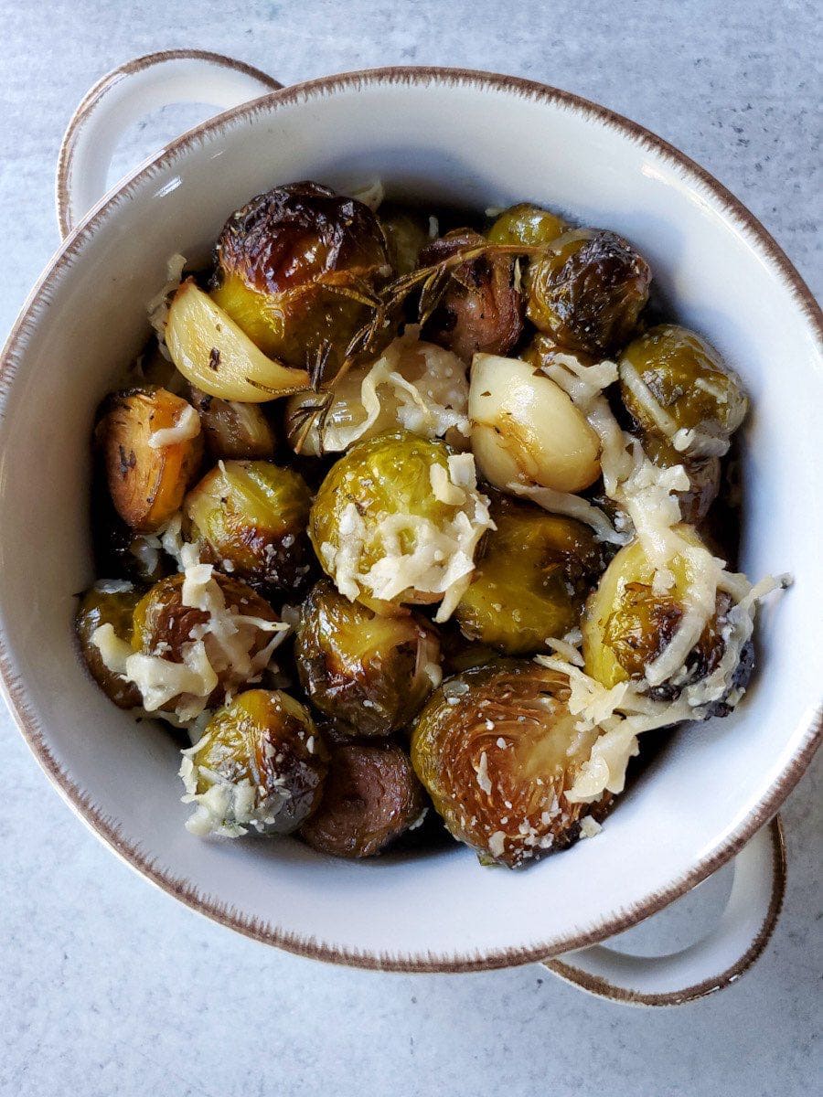 A close up overhead view of a bowl of roasted brussels sprouts, that are slightly blacked in spots, have sprigs of herbs and cloves of garlic showing in the bowl, and a light sprinkle of melted parmesan cheese on top. 