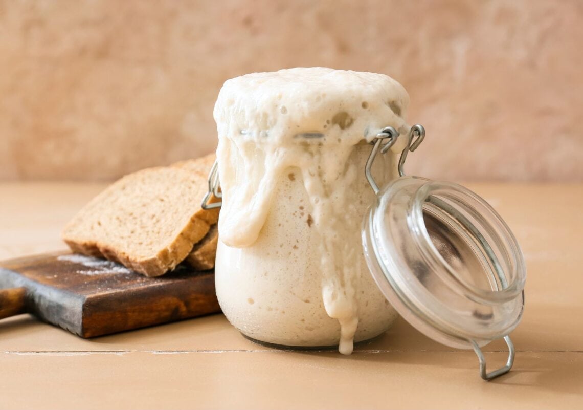 A jar with a flip top lid is overflowing with sourdough starter. Just beyond is a wood cutting board with two slices of bread.